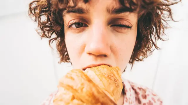 Mulher morde um croissant, piscando um dos olhos. O ângulo exagera o rosto da mulher, o croissant e a mordida.