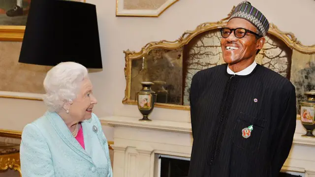 Buhari et la Reine Elizabeth II sont photographiés en conversation, souriant ensembles. La reine porte une veste de costume bleue sur un haut rose, tandis que Buhari porte une veste noire, une chemise blanche et un chapeau à pattes.