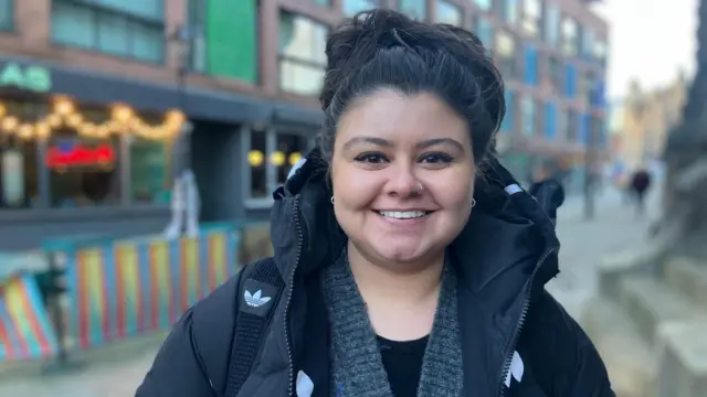 Una mujer con el pelo negro recogido en lo alto de la cabeza sonríe. Lleva un abrigo acolchado. Detrás de ella, Leeds se ve borroso.