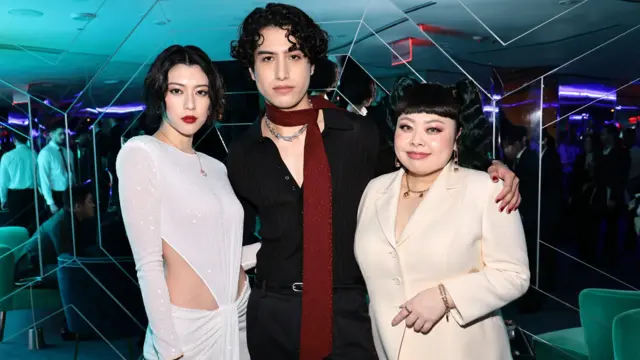 渡辺直美さんが、長身でスリムな男女の隣に立っている。渡辺さんはクリーム色のスーツジャケットと白いロングスカートを着用している。