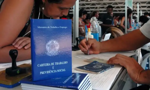 Pilha de carteiras de trabalho em primeiro plano, com mulher sendo atendida por funcionário público para tirar sua carteira ao fundo