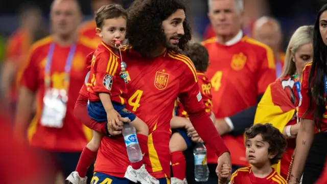 Cucurella con la camiseta roja de España sujeta con una mano a su hijo pequeño y con otra a Mateo