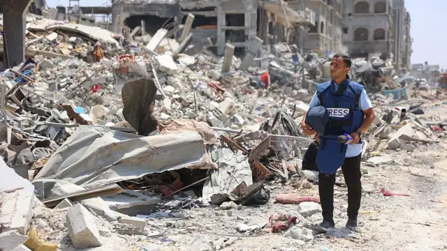 Anas al-Sharif stands wearing a "press" vest and holding a helmet in one hand as he surveys the destruction done to buildings around him in Gaza City. To his left is a huge pile of rubble and debris. 