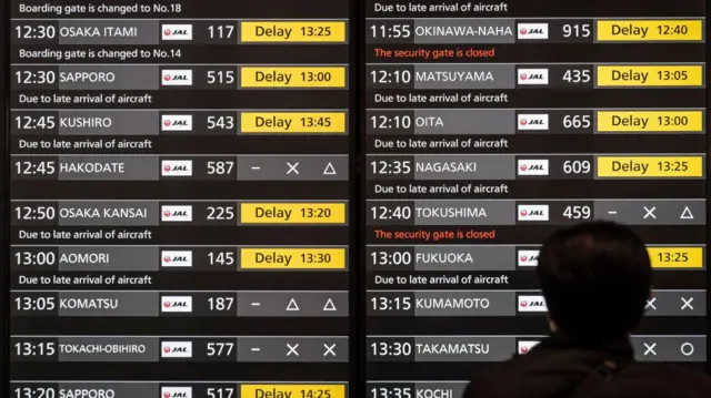 東京・羽田空港の出発ロビーの大きなモニターが、日本航空の運航状況を示している。画面は英語表示で、国内線が軒並み遅延か欠航となっている。右下に、このモニターを眺めている男性の後ろ姿が写っている