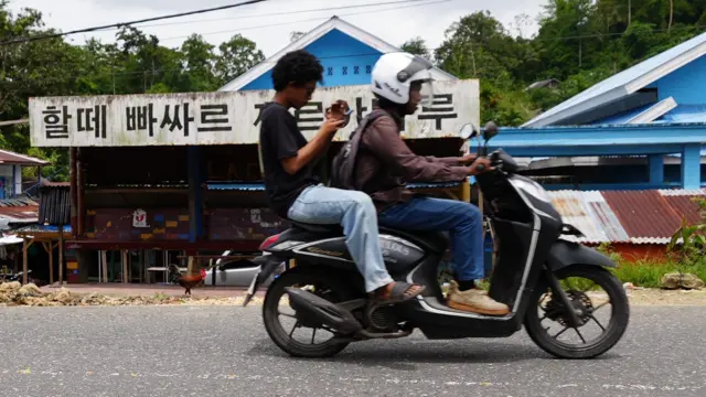 Warga Baubau melintas menaiki sepeda motor di depan Halte Pasar Karya Baru. Palang di halte ditulis menggunakan aksara Hangeul.