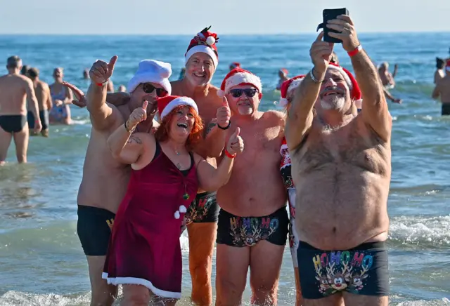 A group of people in the sea wearing santa hats and swimwear, with their thumbs up taking a selfie