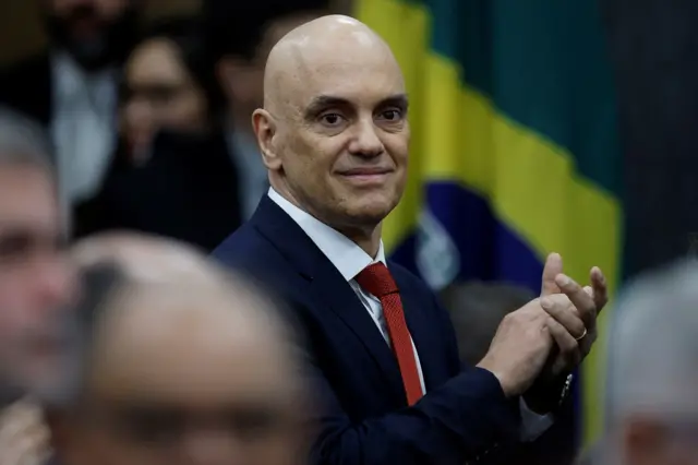 Alexandre de Moraes, um homem branco e calvo, sorrindo em frente a uma bandeira do Brasil 