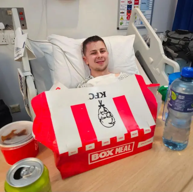 Josh veste um avental hospitalar e está deitado em uma cama de hospital com roupa de cama branca. À sua frente, há uma bandeja com uma garrafa de água, uma caixa de papelão do KFC (nas cores vermelha e branca), um pote de molho do KFC e uma lata de refrigerante aberta.