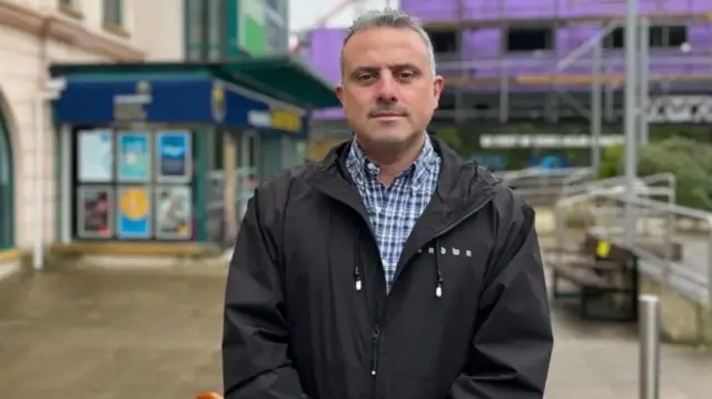 Carl Walker aparece en la foto con chaqueta negra y camisa de cuadros. Detrás de él se ven un supermercado y unos andamios. Tiene el pelo corto y canoso.