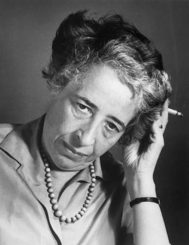 Hannah Arendt con una blusa, un collar, mira hacia abajo y se pone la mano en el cabello. En ella tiene un cigarrillo