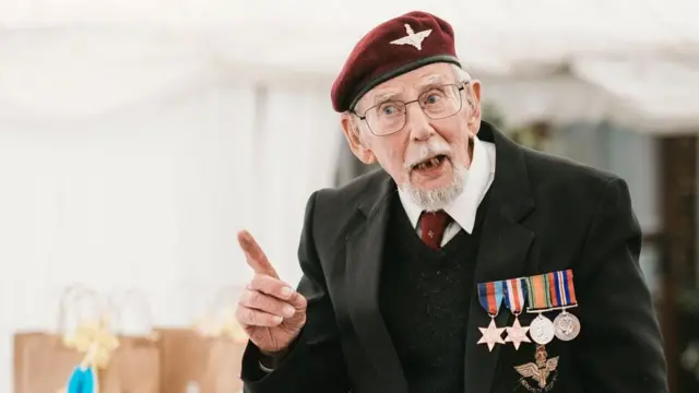 Danny Mason at the 2024 Varsity dinner organised by The Parachute Regimental Association. He has a white goatee beard and is wearing thin black-framed glasses, a black blazer over a black jumper and a white shirt and maroon tie. He has a maroon regimental beret with wings, and on his left breast are four medals. He is speaking and has his right hand raised with his forefinger extended.