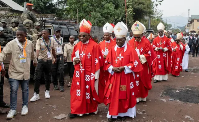 Des évêques catholiques en robes rouges marchent le long d’une rue en terre près de la cathédrale de Goma après avoir célébré la messe suite à la béatification de Floribert Bwana Chui Bin Kositi. Une foule de hauts responsables en costume les suit. Derrière le clerc à gauche, on peut voir un véhicule contenant des soldats rebelles du M23 en uniforme.