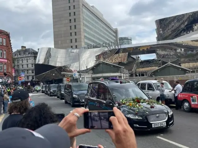 Вереница черных машин едет по городу, на капоте передней машины цветы