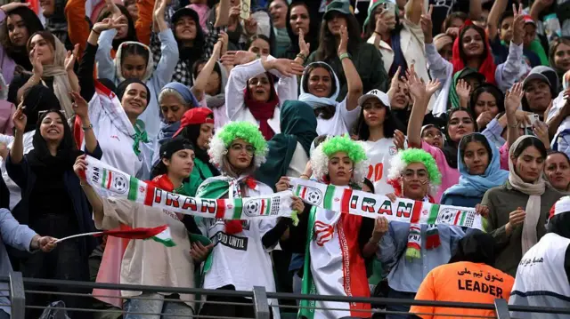 جماهير إيرانية في إحدى مباريات كرة القدم ترتدي النساء شعر مستعار يحمل علم إيران ولافتات عليها اسم إيران