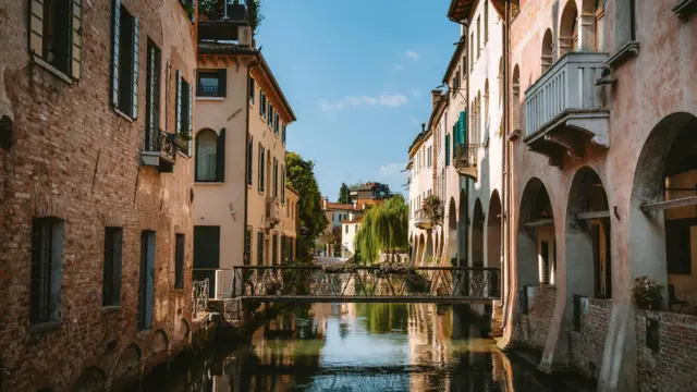 Canalul Cagnan Grande care străbate orașul Treviso, în regiunea Veneto din Italia, cu o pasarelă pietonală care leagă cele două maluri ale orașului. Clădirile din jur se reflectă în apă, în lumina soarelui.