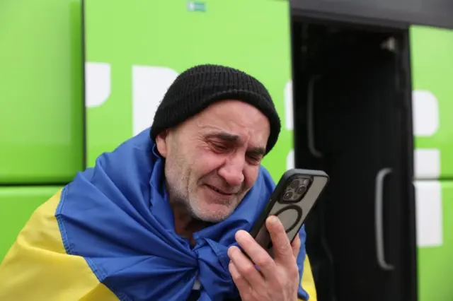 Чоловік, загорнутий в синьо‑жовтий прапор, стоїть біля яскраво‑зеленого автобуса й тримає в руці смартфон, дивлячись на екран зблизька.