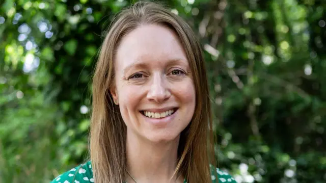 Una foto de Rachel, estilo primer plano. Sonríe a la cámara. Tiene el pelo castaño hasta los hombros y lleva un vestido verde. Detrás de ella hay árboles y arbustos.