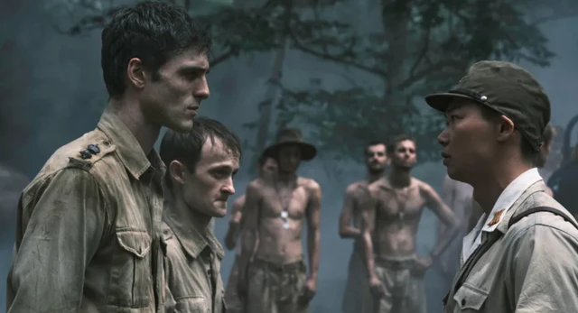 Dos hombres vestidos con un sucio uniforme militar encarados con una mujer de aspecto asiático también de uniforme, aunque limpio. En segundo plano, un grupo de hombres delgados, sucios y con el torso descubierto parecen observarlos. 