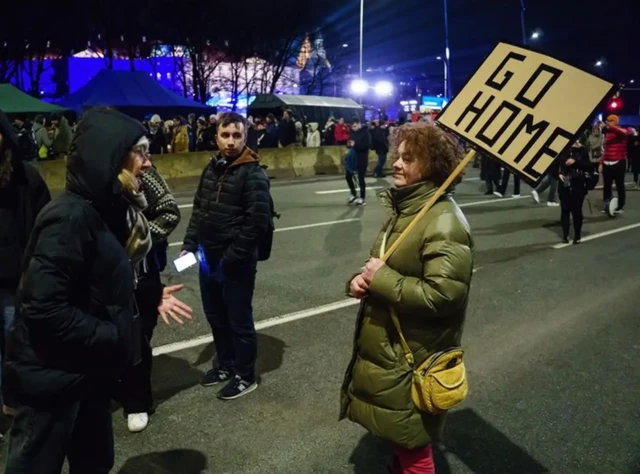 На зображенні – вечірня вулична акція. На широкій проїжджій частині стоїть жінка в довгій зимовій куртці, тримаючи в руках великий картонний плакат із написом "GO HOME". Вона розмовляє з людиною напроти, одягненою в темний одяг із капюшоном.