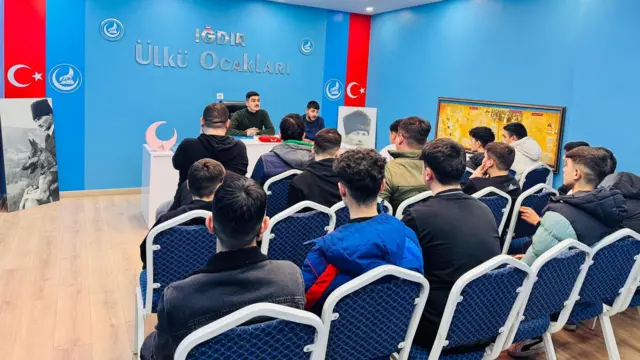 Mavi duvarları olan bir oda, Türk bayrakları var, genç erkekler masada oturan iki kişiyi dinliyor 