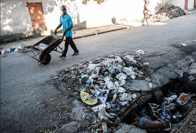 Un hombre con camisa azul pasa empujando una carretilla junto a un montón de basura y una zanja en la que se ve el torso desnudo de una persona muerta en Puerto Príncipe, Haití, el 19 de octubre de 2025.