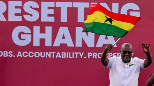 John Mahama se trouve dans le coin droit de la photo, vêtu d'une chemise blanche et agitant un drapeau ghanéen. Derrière lui, une grande affiche proclame : "Reconstruire le Ghana".