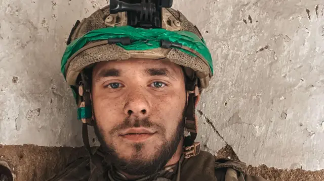 Selfie młodego mężczyzny w ukraińskim mundurze wojskowym. Ma na sobie hełm z aparatem na głowie. Ma krótką brodę i wąsy. Zdjęcie na tle gołej betonowej ściany.