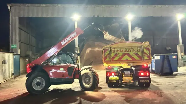 Un vehículo con grúa deposita gravilla en la parte trasera de un camión. Se ve un montón de arena en el almacén trasero.