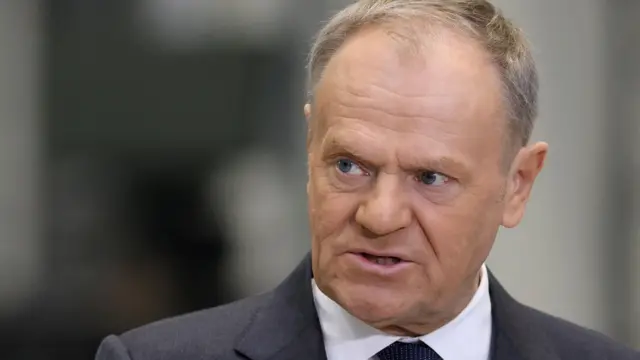 Donald Tusk w ciemnym garniturze i białej koszuli. Stoi w pomieszczeniu o neutralnym, jasnym tle.