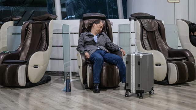 Homem deitado e dormindo em poltrona, aparentemente em aeroporto; ele tem uma mala de rodinhas ao lado 