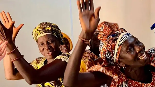 Beberapa perempuan di Gambia mengenakan gaun dan hiasan kepala bermotif warna-warni, bertepuk tangan dan mengangkat tangan.