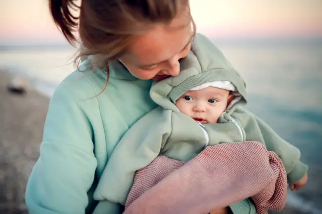 Młoda kobieta w seledynowej bluzie stoi na plaży, trzymając na rękach małe dziecko. Dziecko ma na sobie bluzę w podobnym odcieniu zieleni. Jest owinięte w biało-czerwony koc. Kobieta pochyla głowę w dół, patrząc na nie z uśmiechem. 