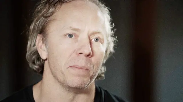 Портрет Грегa Сквайра — мужчины средних лет с длинными седеющими волосами, зачёсанными назад, и серьёзным выражением лица. На нём чёрная футболка, фон тёмный.