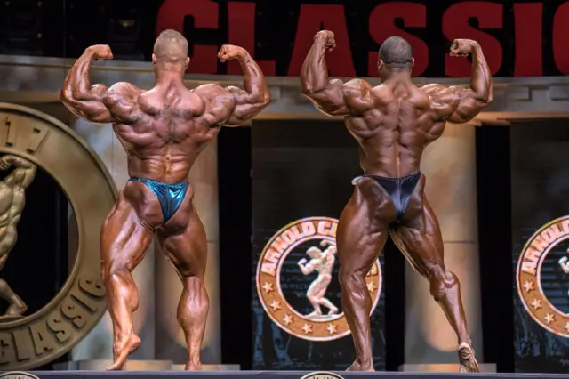 Dois homens musculosos, de costas, exibindo corpos torneados.