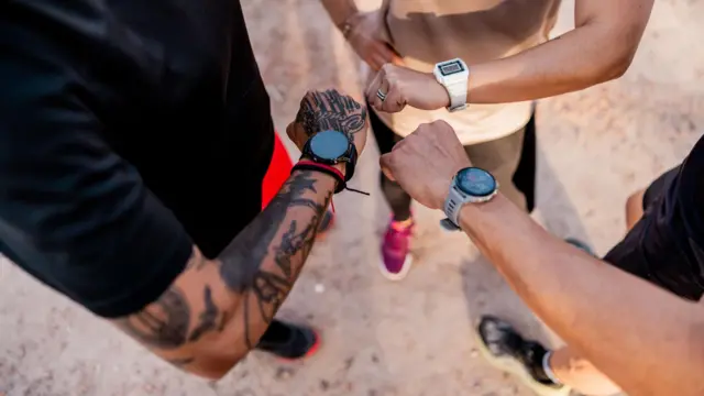 Três corredores olham para seus pulsos com relógios inteligentes acoplados; a imagem foi tirada de cima, com foco em seus braços e pulsos.