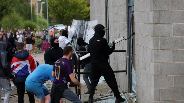 Far-right demonstrators smash windows for The Holiday Inn Express in Rotherham during demonstration. A man dressed in all black with his face covered is at the front.