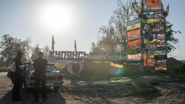 Вказівник Купʼянського району з прапорами, біля нього авто та двоє чоловіків