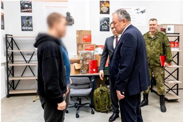 Два студента, заключивших контракт на службу в войсках беспилотных систем, стоят перед ректором вуза в присутствии человека в военной форме и другого официального лица