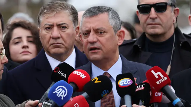 Özgür Özel önünde gazetecilerin uzattığı mikrofonlara konuşuyor. Arkasında CHP Ankara milletvekili Murat Emir ve güneş gözlüklü bir koruma var. 