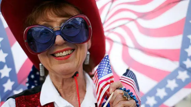 Una mujer con una sombrilla con los colores de la bandera de Estados Unidos, vestida con un sombrero estilo vaquero rojo, chaleco de lentejuelas rojo y azul, camisa blanca y unos lentes azules y rojos. Lleva en su mano dos pequeñas banderas de Estados Unidos.