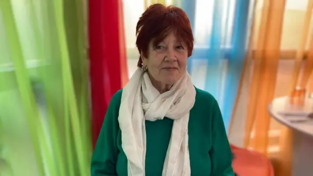 Shirley Heard, sentada en un café, con un jersey verde y una bufanda color crema. Tiene el pelo corto y pelirrojo y pendientes de plata. Detrás de ella hay largas cortinas de colores: verde, rojo, azul y naranja. Mira a la cámara.