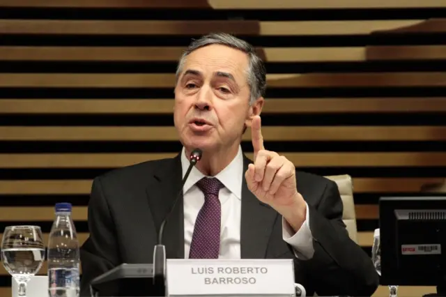 Barroso gesticulando enquanto fala, sentado, em salão; à sua frente, placa com seu nome