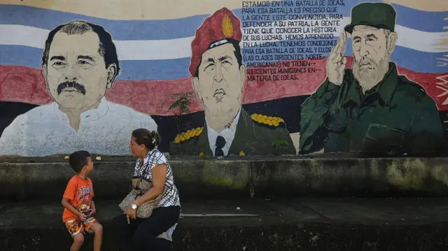 Mulher e criança conversam em frente a um mural em Manágua, na Nicarágua, retratando Daniel Ortega, Hugo Chávez e Fidel Castro