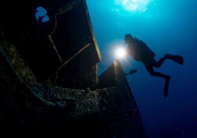 A diver shines a flashlight under water at a wreck.