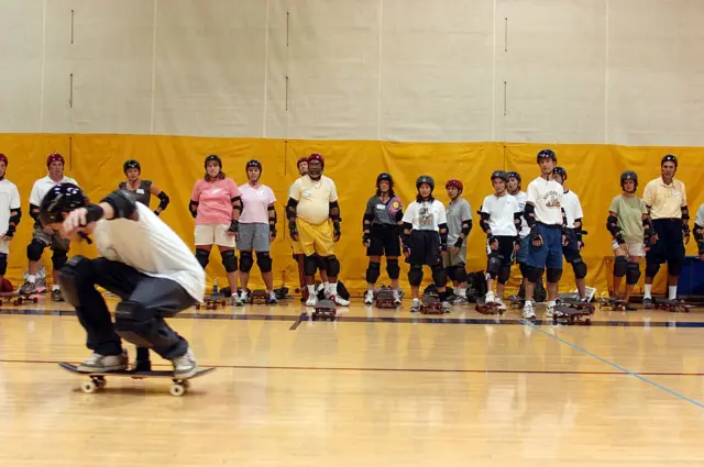 Grupo de pessoas aprendendo a andar de skate. Uma pessoa no centro faz manobras enquanto os outros assistem