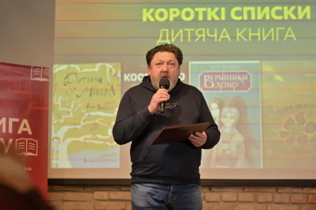 Іван Андрусяк на сцені розповідає про свою книгу. В руках у нього відзнака