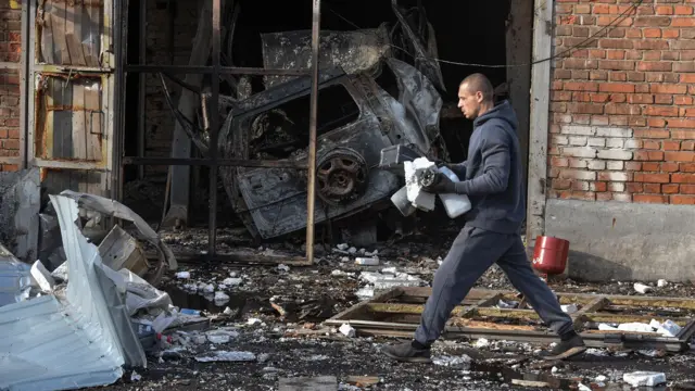 Mężczyzna idzie ulicą, na której leży gruz, obok spalony samochód, w zniszczonym warsztacie mechanika samochodowego w Dnieprze 