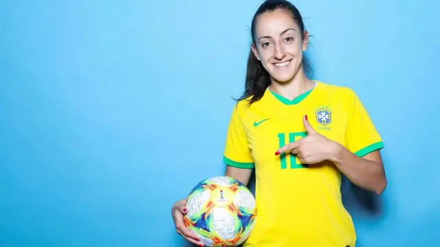 Luana Bertolucci veste a camisa amarela e verde do Brasil e segura uma bola de futebol. Ela está com o cabelo preso e sorri para a câmera