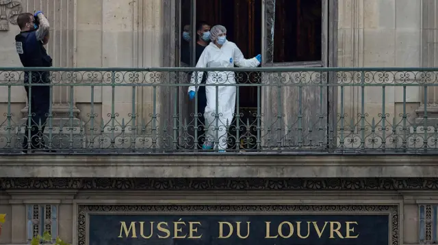 Osoba w białym kombinezonie stoi w otwartych drzwiach na balkonie, a lewej strony mężczyzna wygląda jakby robił zdjęcie