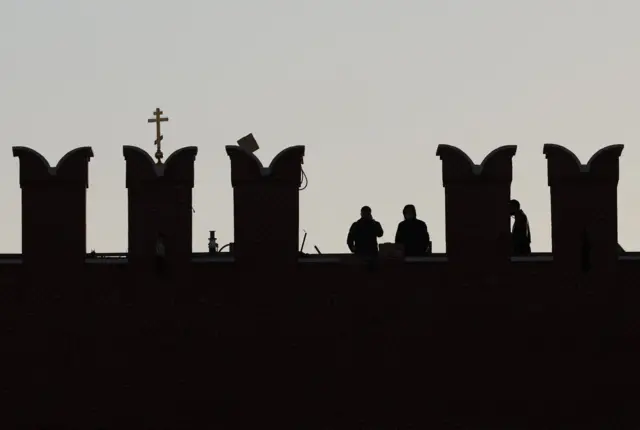 На фоне светло-серого неба видны очертания зубцов крепостной стены Кремля. Изображено пять зубцов. Из-за второго виден крест с навершием и косой перекладиной внизу. Между зубцами видны три человеческих силуэта.